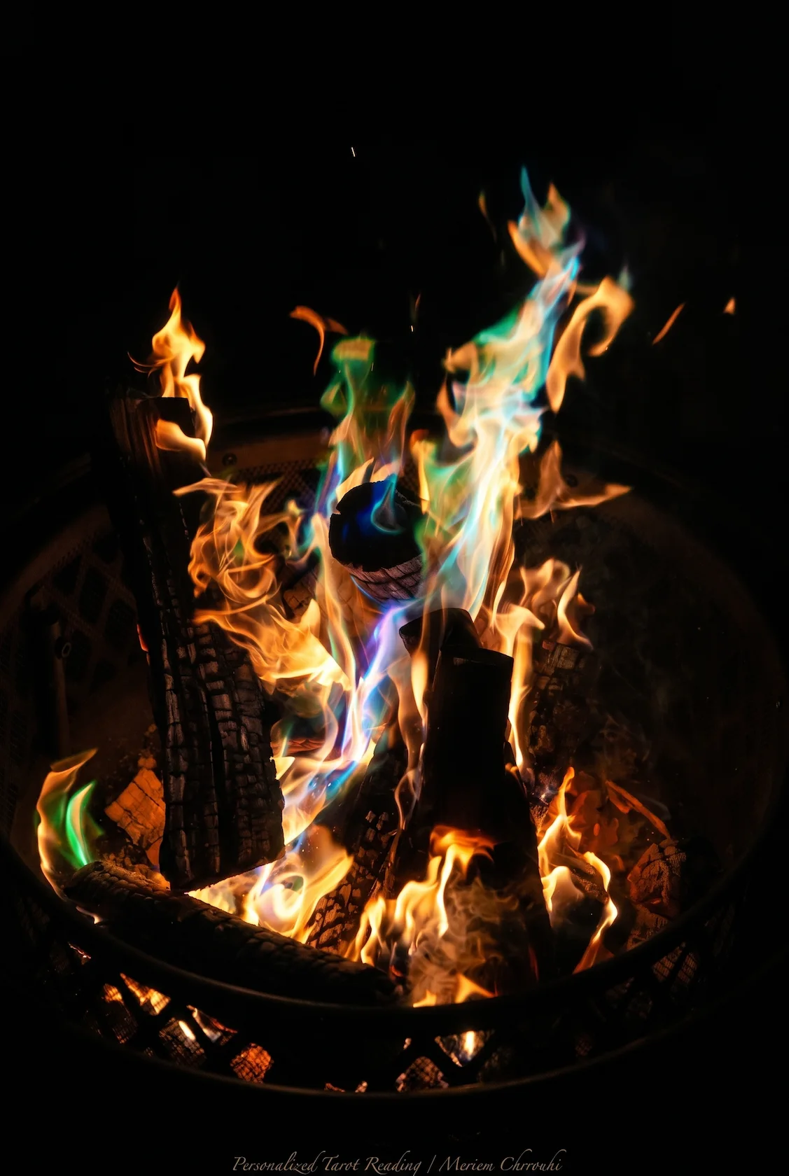 Gold and blue-green flames rising from logs in a dark metal fire basket.