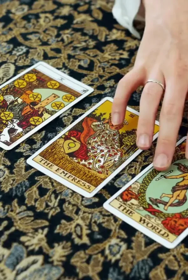 Hand resting beside The Empress, The World and Ten of Pentacles tarot cards on patterned fabric.